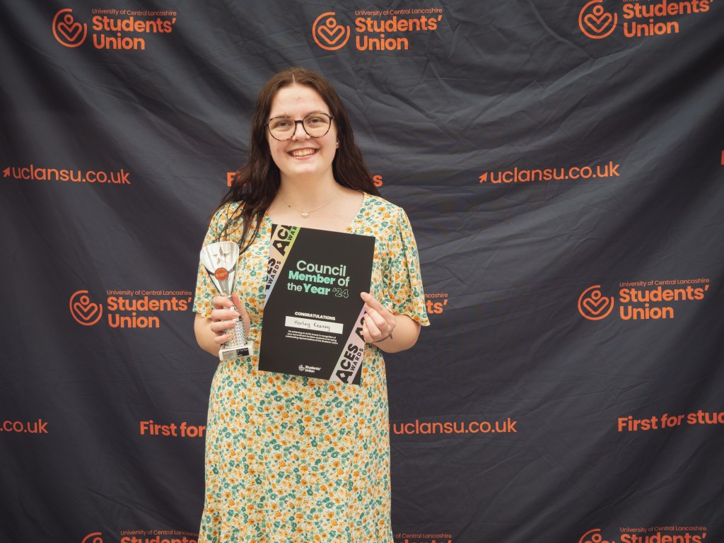 Person stands smiling in front of a Students’ Union backdrop, holding a certificate and a trophy.