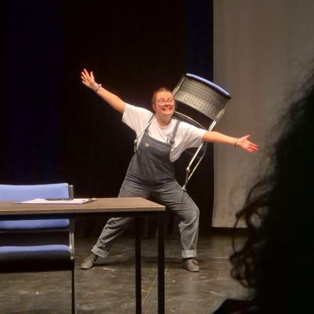 Performer stands on stage with arms outstretched, smiling, while wearing a chair strapped to her back beside a table.