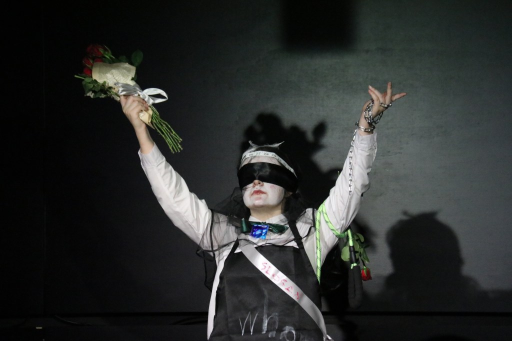 Performer wearing a blindfold raises a bouquet of flowers and one arm upward on a dark stage, as part of a live performance.