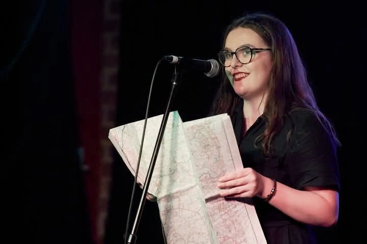 Performer stands at a microphone holding an unfolded map, smiling as she reads during a live spoken performance.