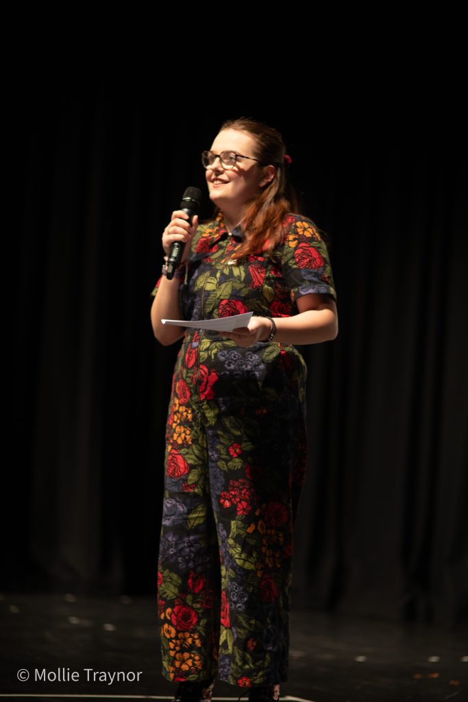 Speaker stands on stage holding a microphone and notes, wearing a floral jumpsuit, addressing an audience.